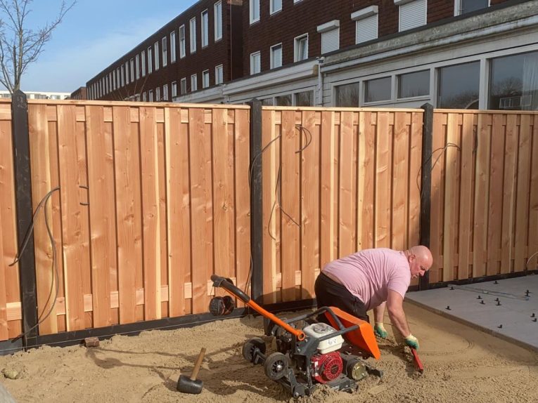 Hovenier met roze shirt op zijn knieen aan het werk in het zand in de tuin